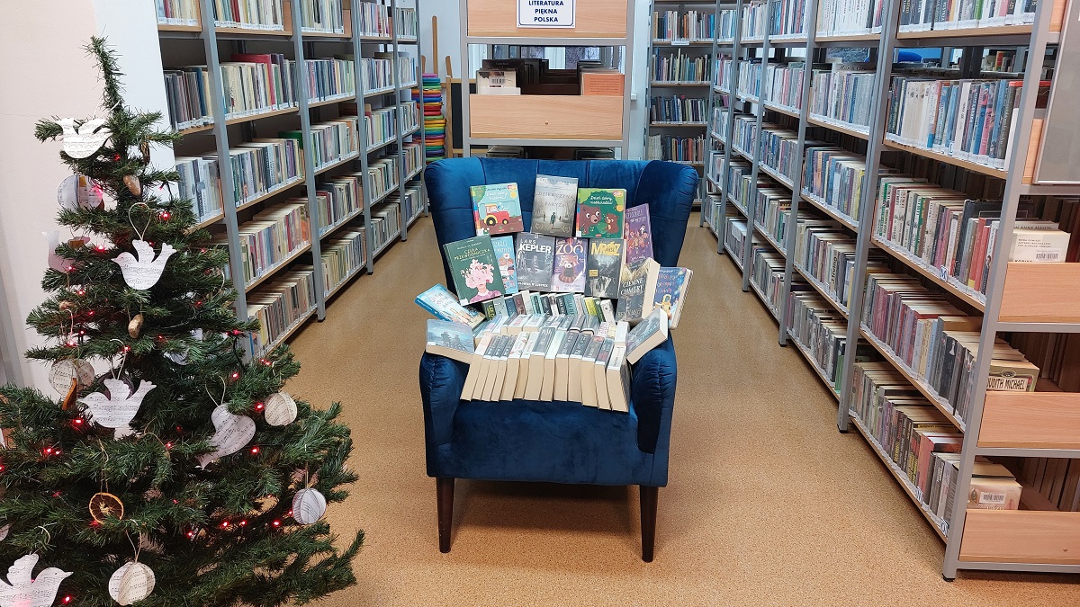 Sala wypożyczalni biblioteki. Pomiędzy regałami z ksziążkami stoi fotel na którym leżą książki. Z lewej strony fotela stoi ubrana choinka.  