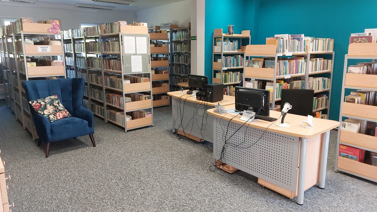 Pomieszczenie biblioteczne. Na środku stoją dwa biurka. Pod ścianami stoją regały z książkami